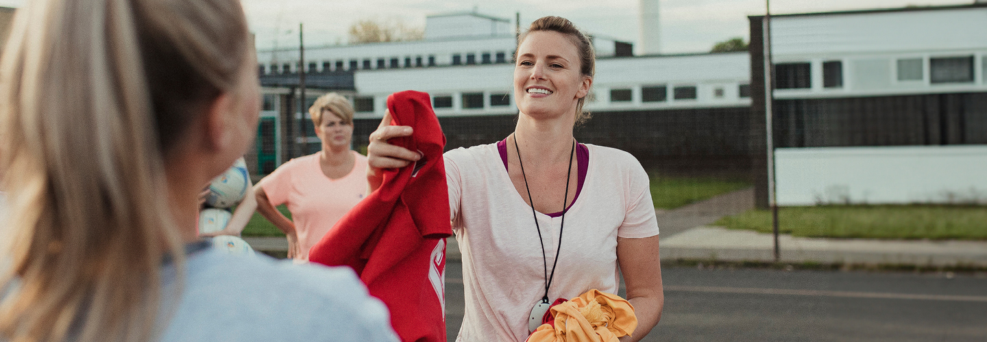 A blonde netball coach smiles as she hands out bibs to her team. She wears a pale pink t-shirt and a whistle around her neck